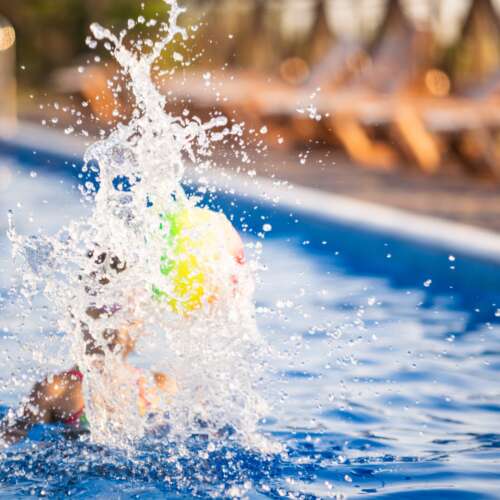 A big slash in a swimming pool by a young girl in a bright leopard swimsuit, swims with a bright colored inflatable ball in a deep blue pool with clear transparent water on a warm sunny summer evening